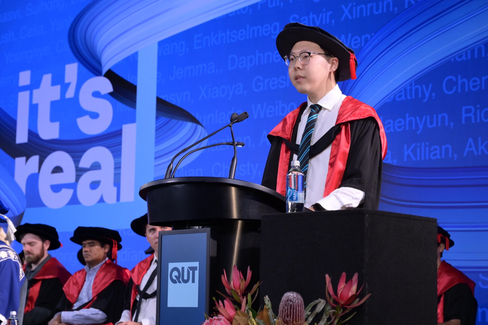Zhipeng He speaking at the lectern in doctoral regalia at the QUT graduation ceremony, Star Brisbane